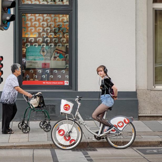 Junges Mädchen auf einem Fahrrad, daneben eine alte Dame mit einem Rollator.